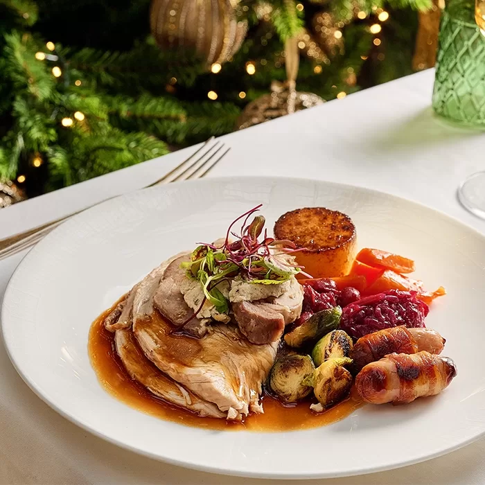 A festive plate with sliced roast turkey, gravy, sprouts, bacon-wrapped sausage, carrot, red cabbage, a round stuffing, and a garnish, set on a white table with Christmas decorations in the background.