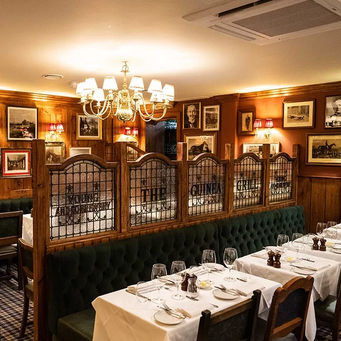 A cozy restaurant interior with white tablecloths, set tables, dark green tufted seating, wooden dividers with stained glass, warm lighting from chandeliers, and framed art on wood-paneled walls.