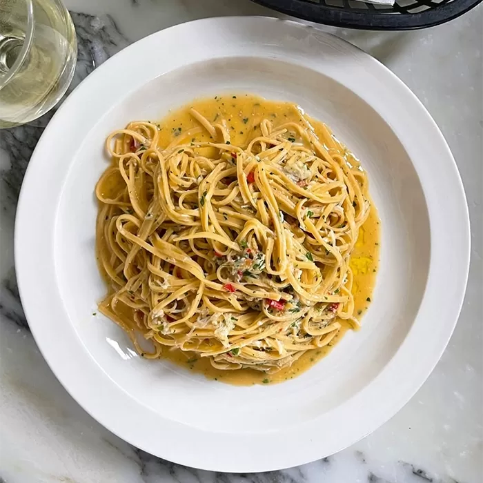 A plate of spaghetti with a creamy orange sauce, herbs, and small bits of vegetables, served on a white plate atop a marble table. A glass of white wine is partially visible in the corner.