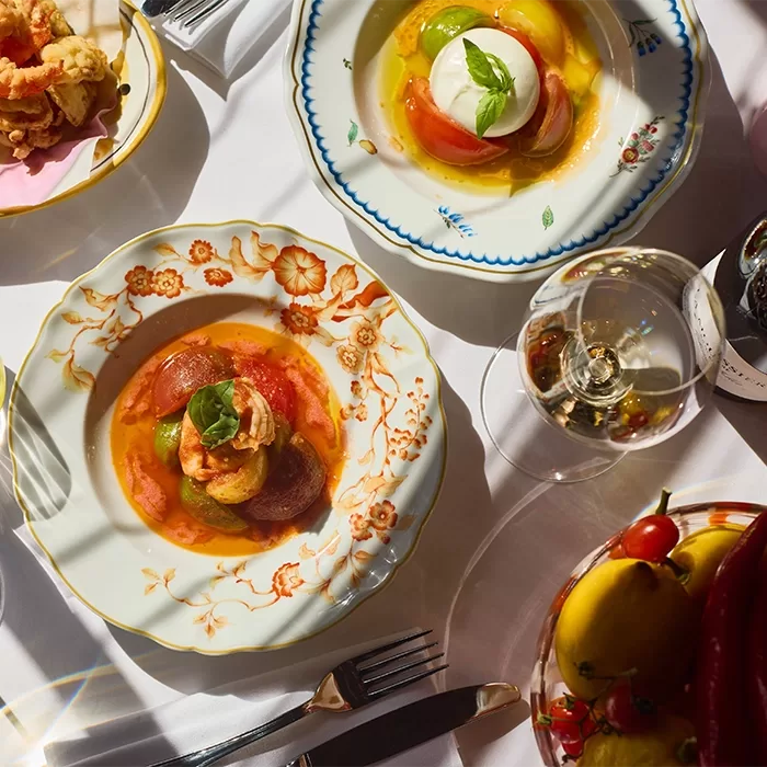 Plates of colorful tomato and burrata salads on ornate dishes are set on a white tablecloth, with a glass of white wine, utensils, and a small bowl of tomatoes visible in warm, natural light.
