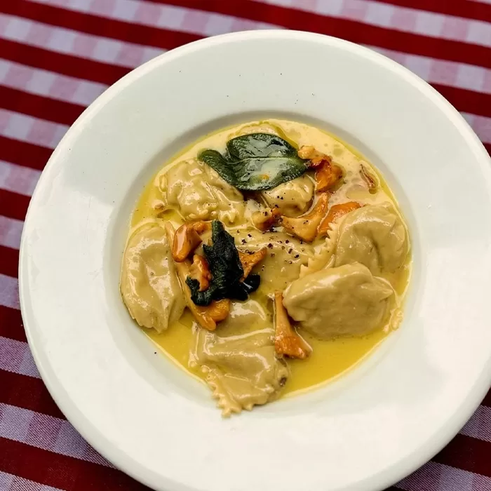 A white plate with four ravioli in a creamy yellow sauce, garnished with fresh herbs and mushrooms, sits on a red and white checkered tablecloth.