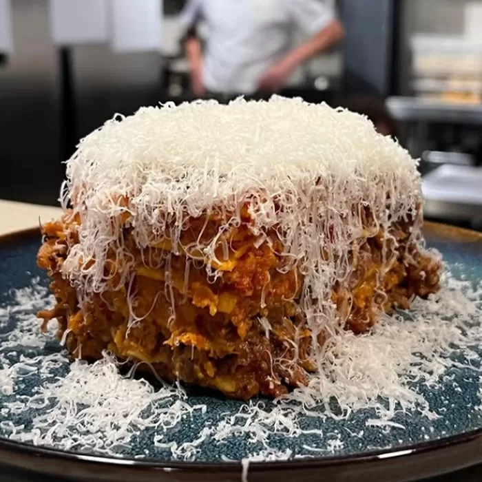 A thick slice of lasagna topped with a large mound of finely grated cheese sits on a blue plate; a person in chef attire is blurred in the background.