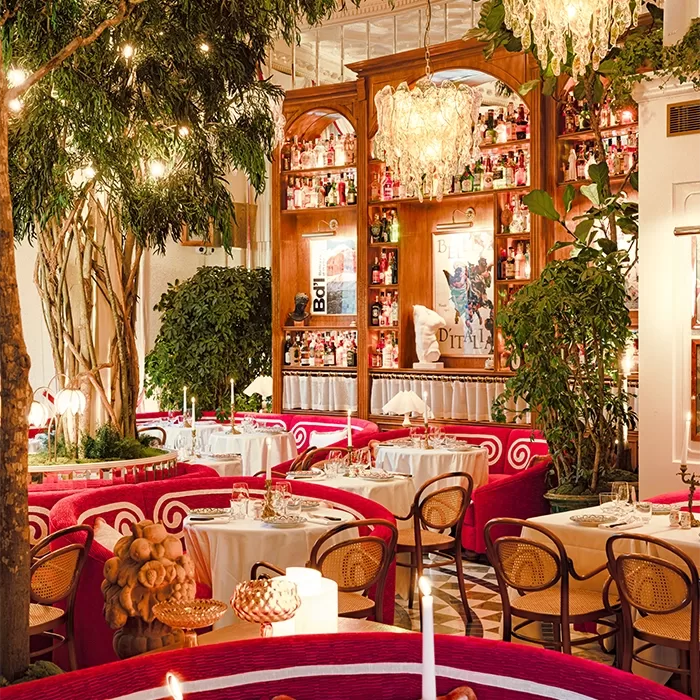 A vibrant, elegant restaurant with red cushioned seating, round tables set for dining, lush indoor greenery, chandeliers, and a large wooden bar lined with bottles and illuminated by warm lighting.