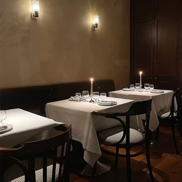 Three tables with white tablecloths are set for dining in a dimly lit restaurant, each with a lit candle and glassware. Dark wooden chairs and soft wall lighting create a cozy, intimate atmosphere.