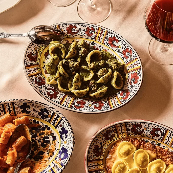 Three colorful patterned plates on a table: one with pasta and green pesto, one with red sauce pasta, and one with ravioli in sauce. Two wine glasses are partly visible at the top.