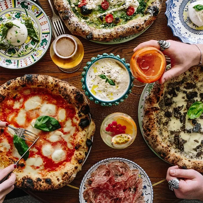 A top-down view of a wooden table with assorted pizzas, drinks, burrata, dip, and cured meats. Two hands are reaching in—one cutting pizza, another holding an orange drink. The scene is vibrant and inviting.