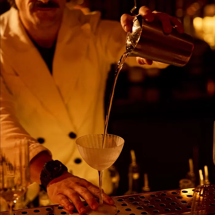 A bartender in a white jacket pours a cocktail from a metal shaker into a coupe glass at a dimly lit bar.