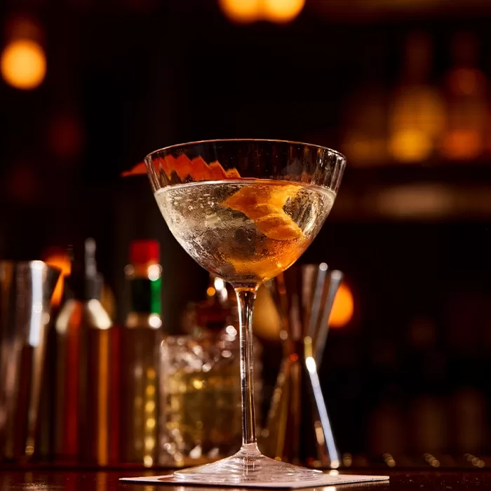 A cocktail in a stemmed glass garnished with a citrus twist sits on a bar, with blurred bottles and bar tools in the background under warm lighting.