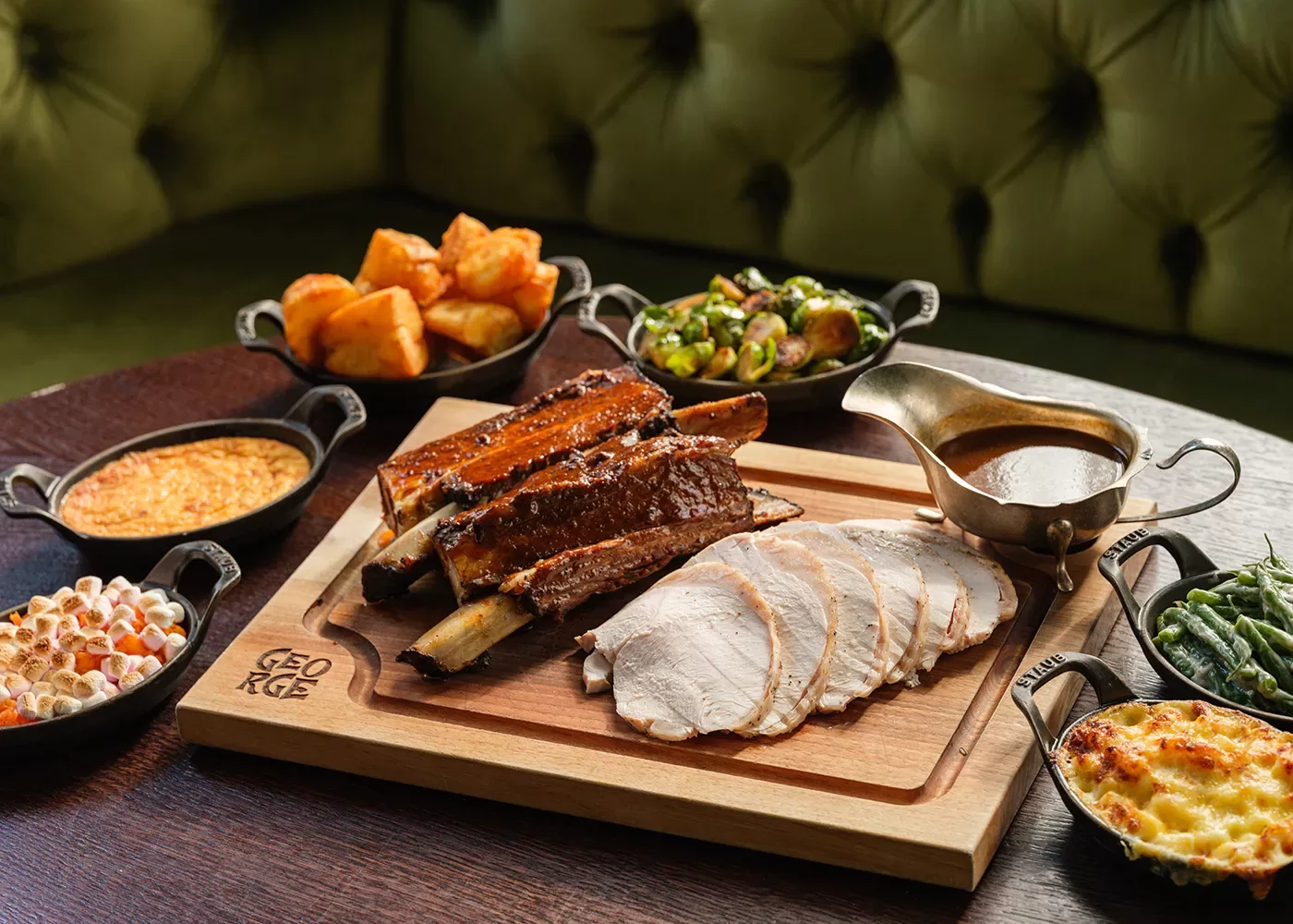 A wooden board with sliced roast meat and glazed ribs, surrounded by small dishes of roasted potatoes, Brussels sprouts, creamy corn, green beans, macaroni and cheese, and brown gravy.