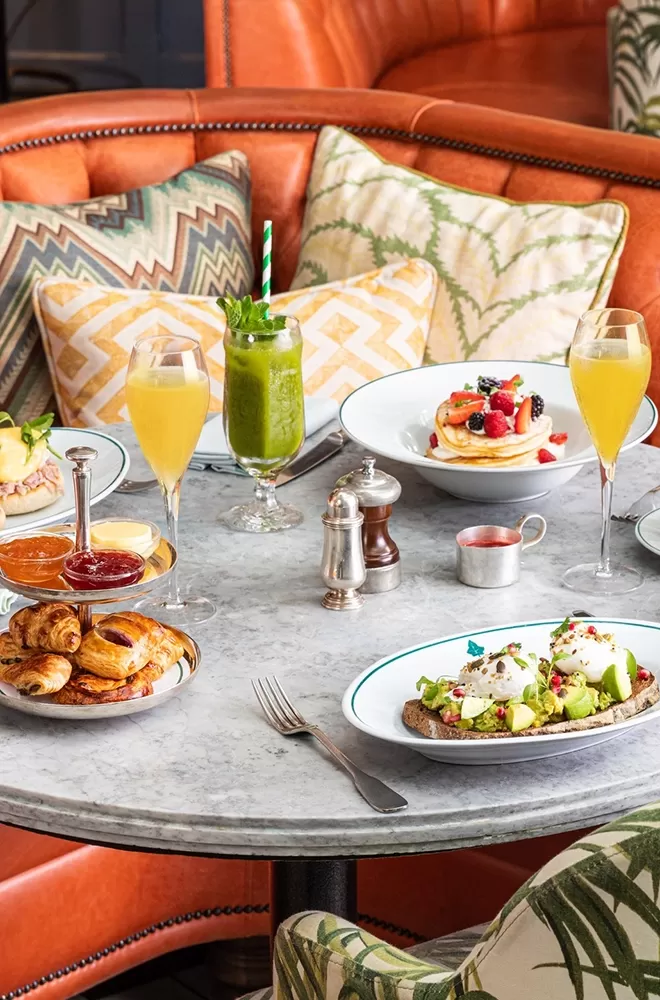A round marble table set for brunch with two glasses of orange juice, a green smoothie, pancakes topped with berries, avocado toast with eggs, a plate of mini pastries, various jams, and a silver pepper grinder. Decor includes patterned cushions at The Ivy, London.