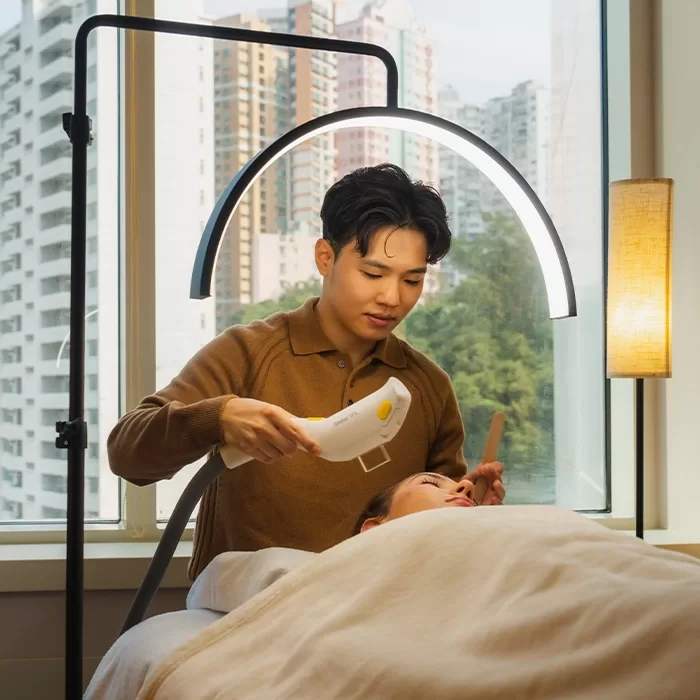 A person uses a handheld device on a clients face in a spa or clinic setting, with a bright arched lamp overhead and large windows showing tall buildings and greenery outside.