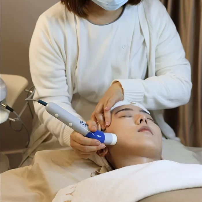 A person receives a facial treatment while lying down, with a clinician using a handheld skincare device on their face. The clinician wears a white coat and mask. The patient’s eyes are closed and a towel covers their hair.
