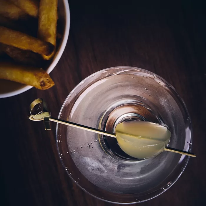 A top-down view of a martini garnished with a green olive on a cocktail pick, next to a bowl of fries on a dark wooden surface.