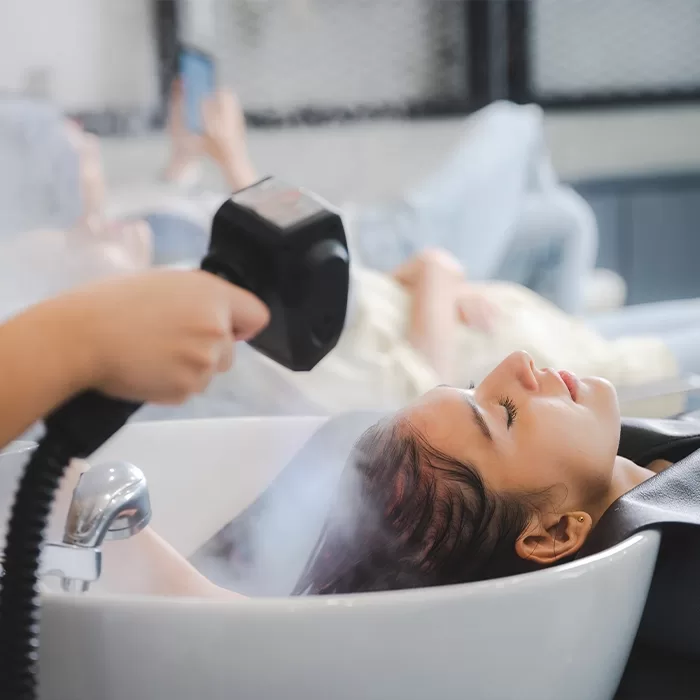 A woman relaxes with her eyes closed at a salon, having her hair steamed by a stylist while reclining at a sink.