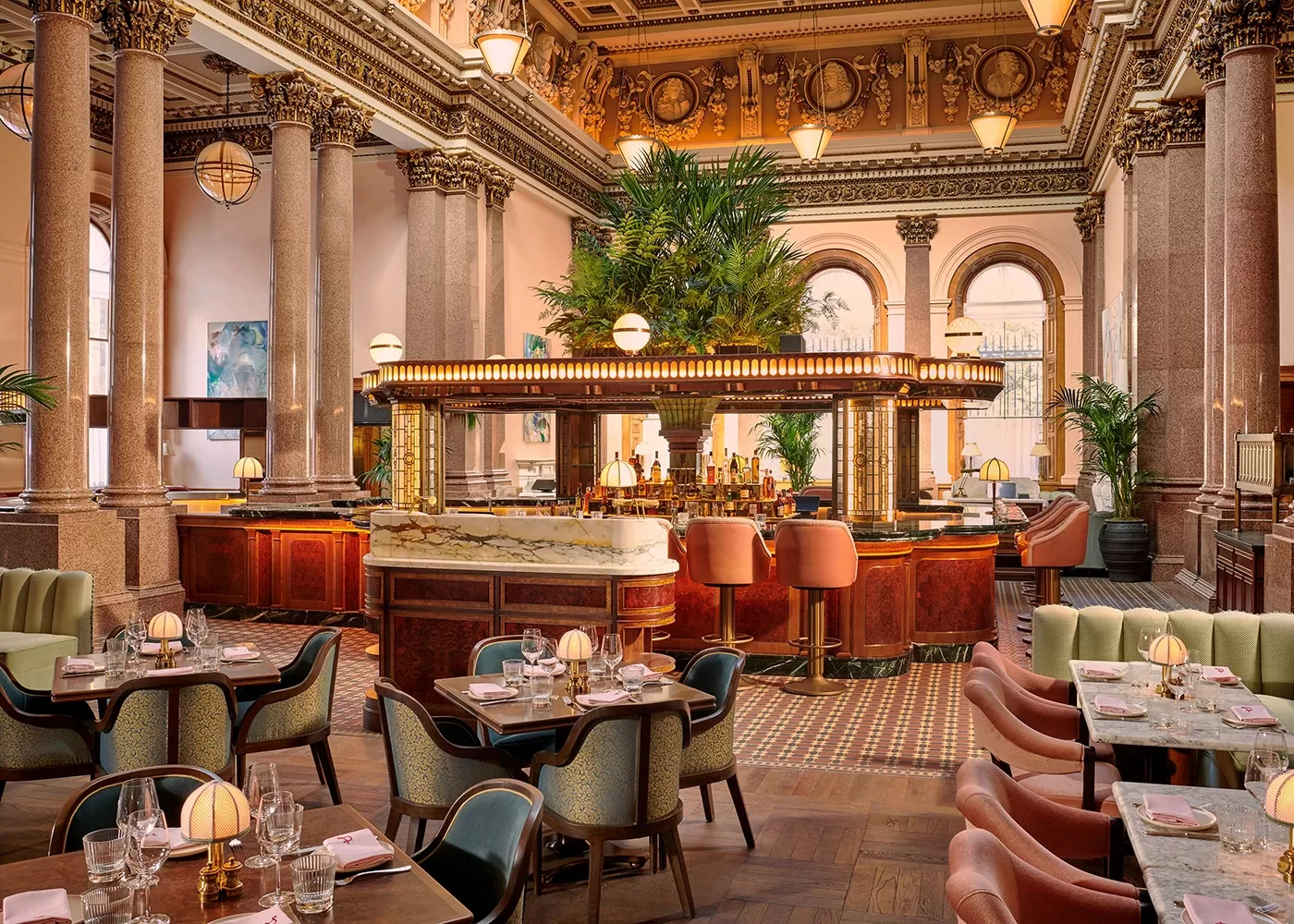 Elegant restaurant with high ornate ceilings, large arched windows, marble columns, and a central bar surrounded by pink and green upholstered chairs and lush green plants. Tables are set for dining with glassware and napkins.