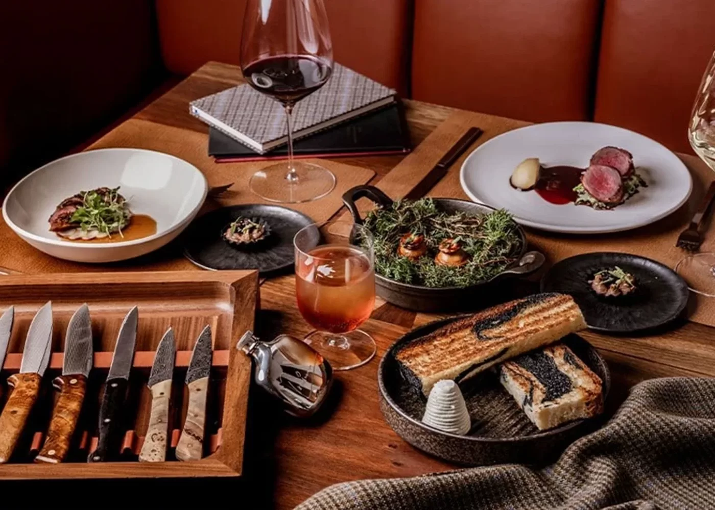A wooden table set with gourmet dishes, including steak, greens, grilled bread with butter, small appetizers, glasses of red and orange drinks, cutlery, and notebooks in a cozy restaurant setting.