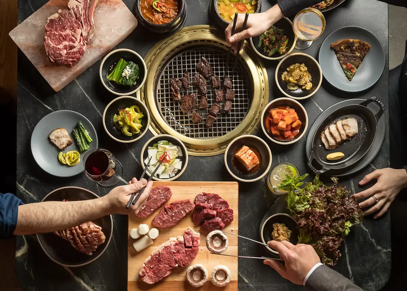 Top view of a Korean BBQ meal, featuring a grill with meat cooking, surrounded by side dishes, raw meats, and hands holding chopsticks and cutting meat. There are plates of vegetables, sauces, and drinks on a dark table.