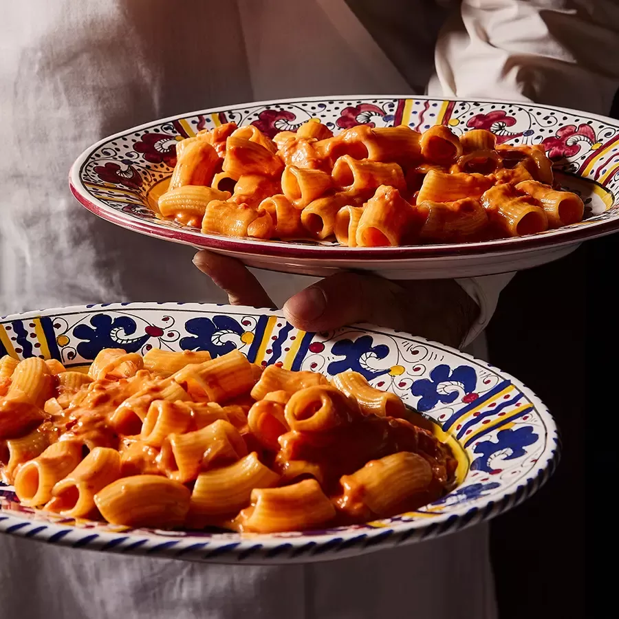 Two colorful, patterned plates filled with rigatoni pasta in a rich, red tomato sauce are being held by a person in a white shirt.