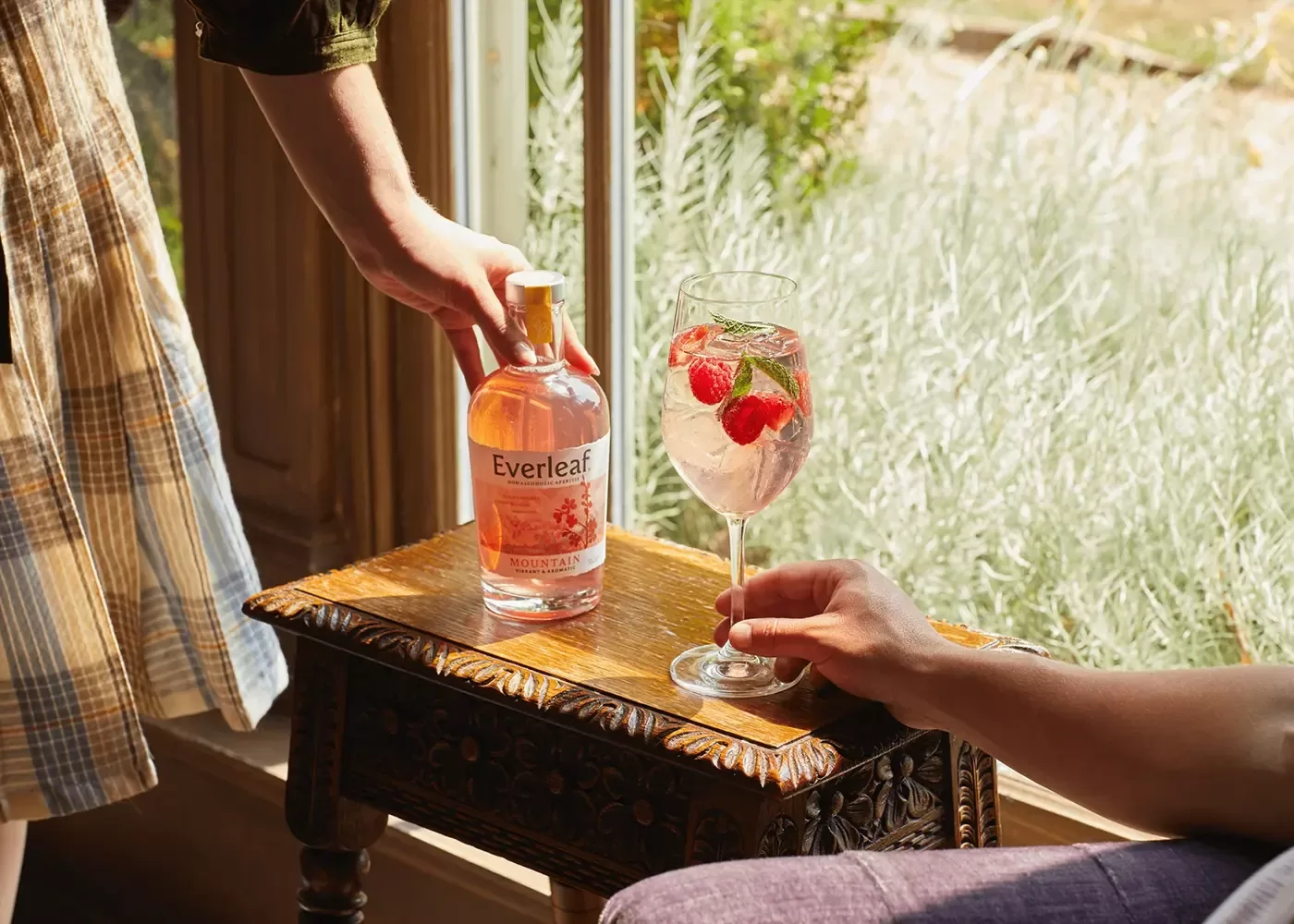 Women holding a bottle of Everleaf Mountain apéritif on a side table with a glass of drink with strawberry garnish next to a window.