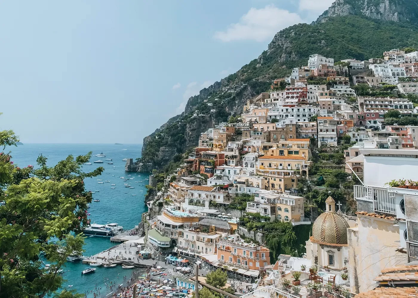Colorful buildings stacked on a steep hillside overlooking a sparkling blue sea with boats, lush greenery, and a crowded beach below; mountains rise in the background under a clear, sunny sky.
