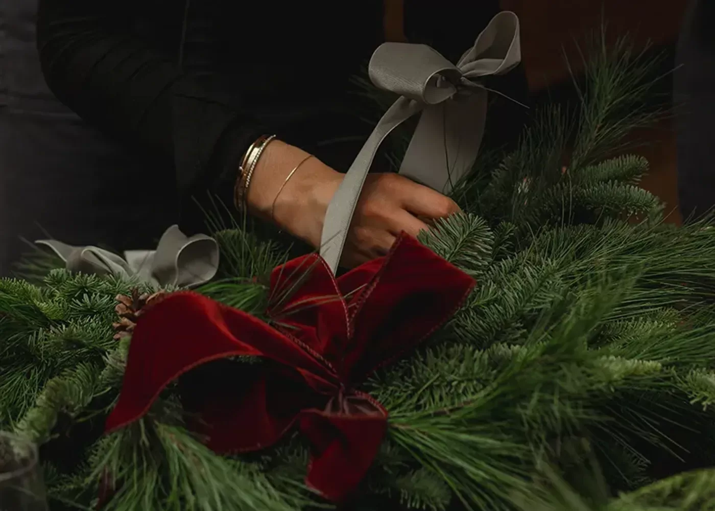 A person’s hands are decorating a green pine wreath with a large red velvet bow and a gray ribbon. The person is wearing a black sleeve and a silver bracelet.