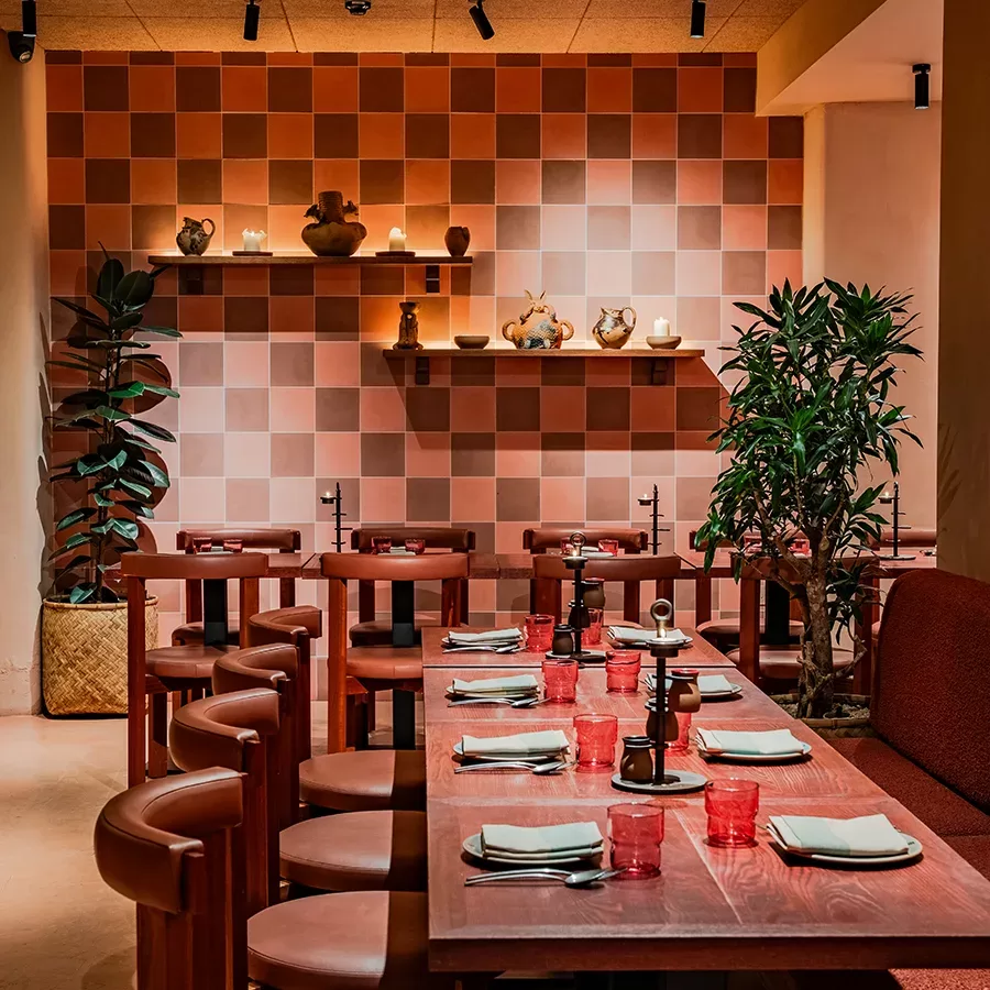 A cozy restaurant interior with pink square-tiled walls, wooden tables and chairs set for dining, red glassware, potted plants, and shelves displaying pottery and lit candles. Warm lighting creates an inviting atmosphere.