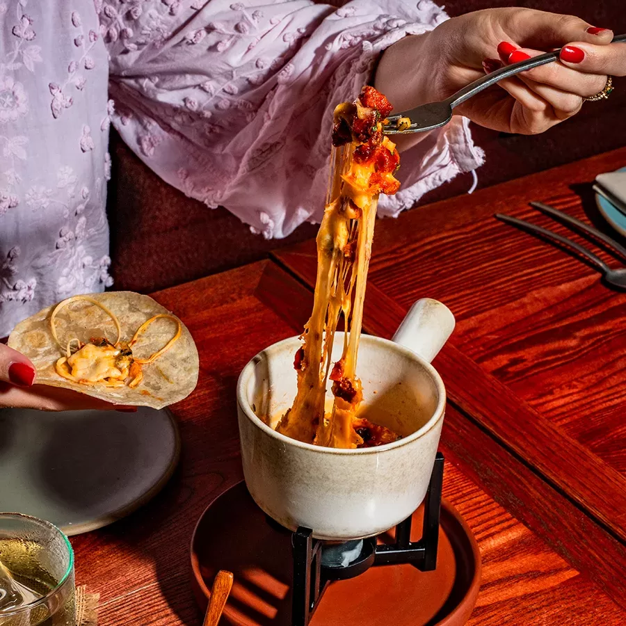 A person in a pink, ruffled sleeve scoops melty cheese with chorizo from a cup using a fork, stretching the cheese onto a tortilla over a wood table set with utensils and a drink.