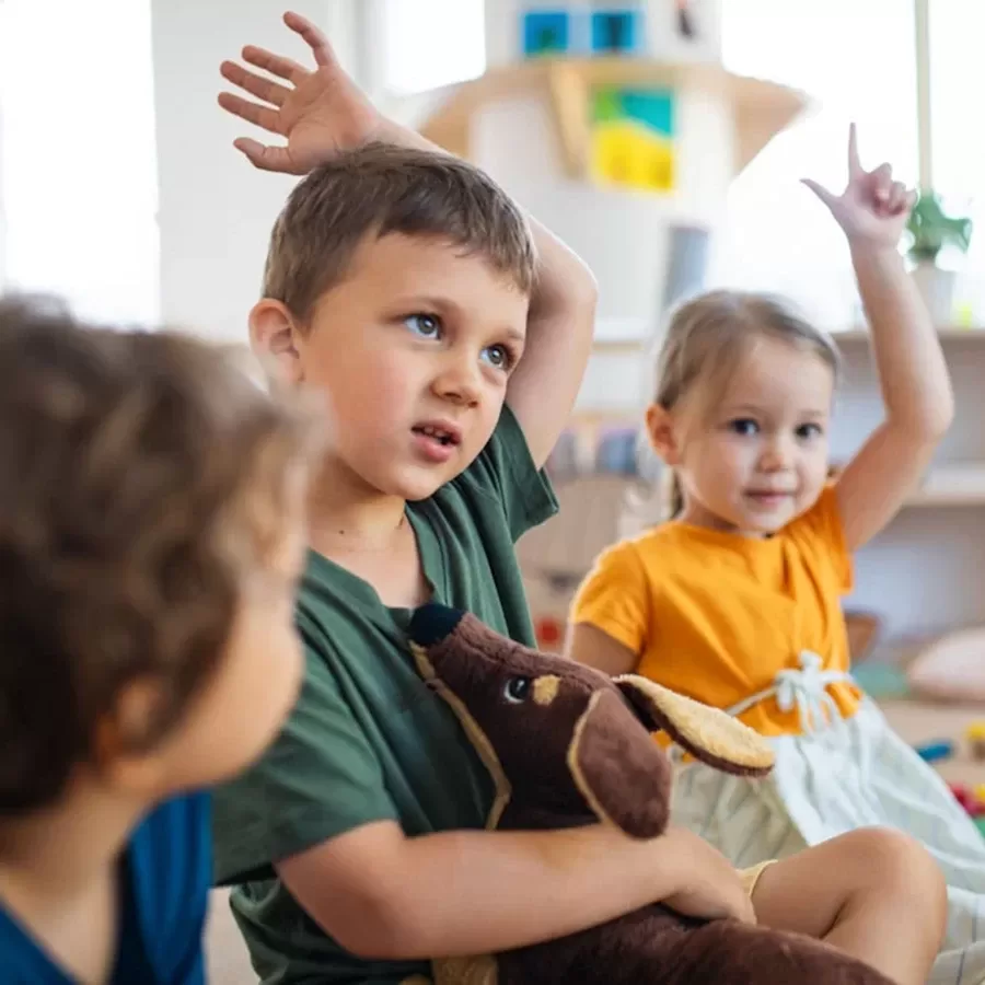 Three young children sit indoors; two have their hands raised. The boy in the center holds a plush dog toy and looks up, while the girl in orange also raises her hand and smiles. The scene appears to be in a classroom or playroom.