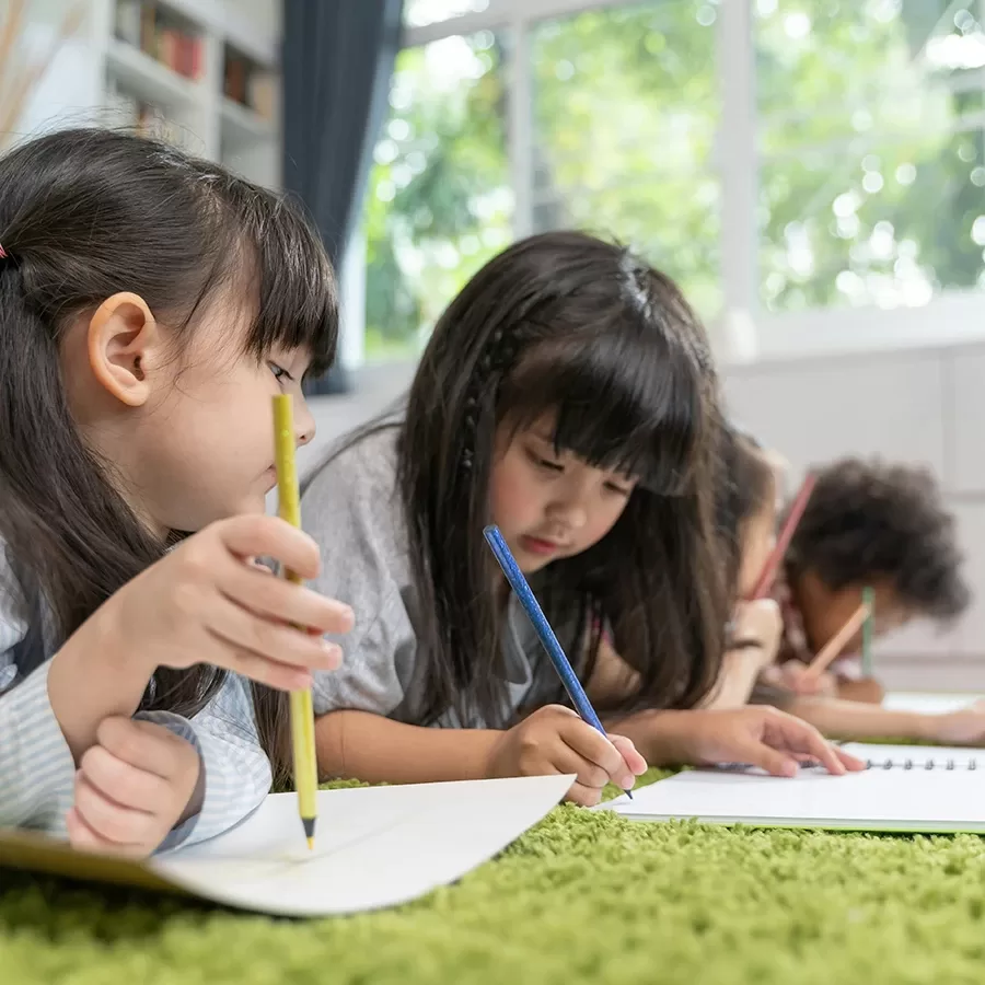 Four young children lie on a green carpet, drawing or writing in notebooks with colored pencils. They are focused on their work, and large windows in the background let in natural light.