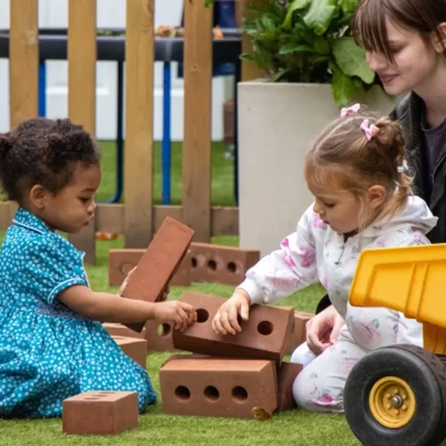 Two young girls play with toy bricks on grass while a woman, possibly a teacher or caregiver, watches them and smiles. A yellow toy truck is nearby, and a wooden fence and plants are in the background.