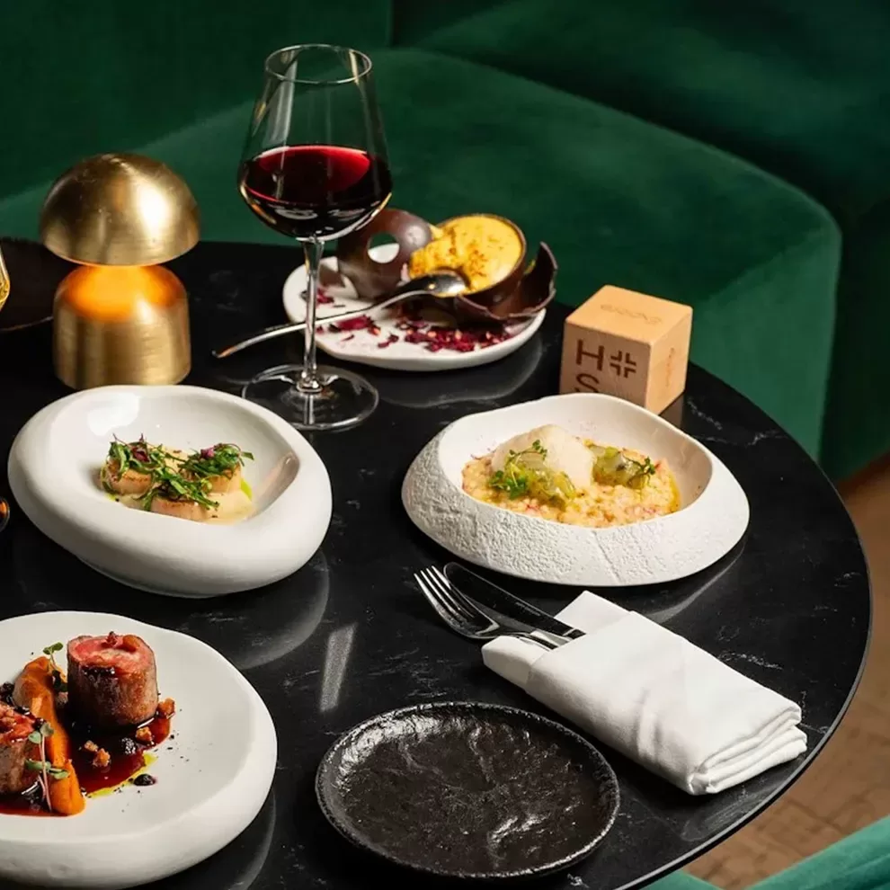 Food and drink served on round dining table with green velour sofa interior at Hithe + Seek, London