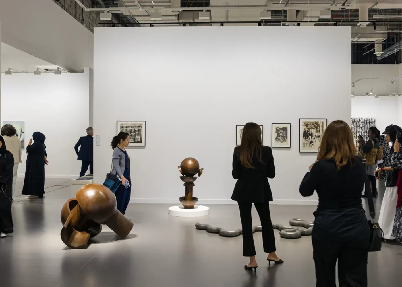 People observe sculptures and framed art on display in a modern gallery. The sculptures are abstract and metallic, and the gallery has bright lighting with a plain white wall as the backdrop.