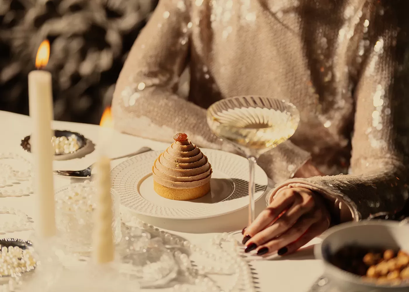 A person in a sparkling, sequined top sits at a table set with a dessert, a coupe glass of white wine, candles, and elegant tableware, creating a festive and luxurious dining atmosphere.