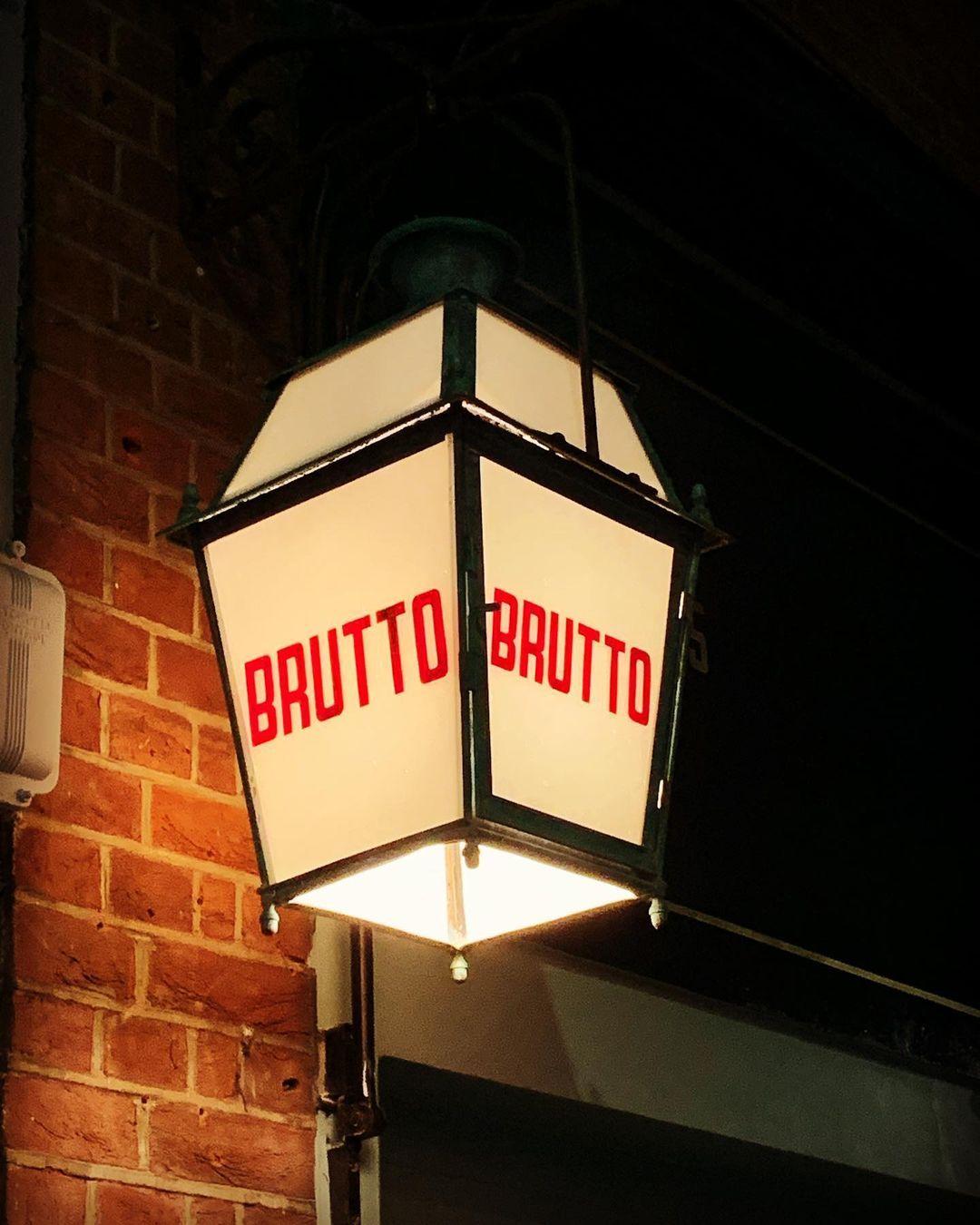 A vintage-style street lamp with the word BRUTTO in red letters on two sides, mounted on a brick wall. The lamp is illuminated, casting a warm glow against the dark background at Brutto, Farringdon.