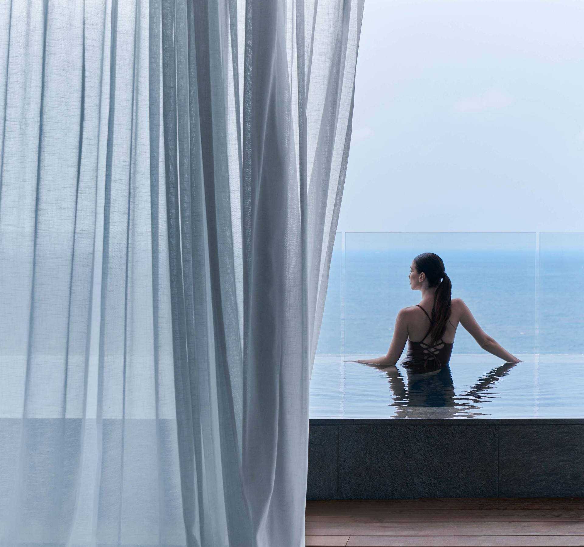 A woman in a swimsuit sits on the edge of an infinity pool overlooking the ocean. She is gazing at the sea, with her back turned to the camera. Sheer curtains partially frame the scene on the left side at SHA Wellness Clinic Mexico, Mexico.
