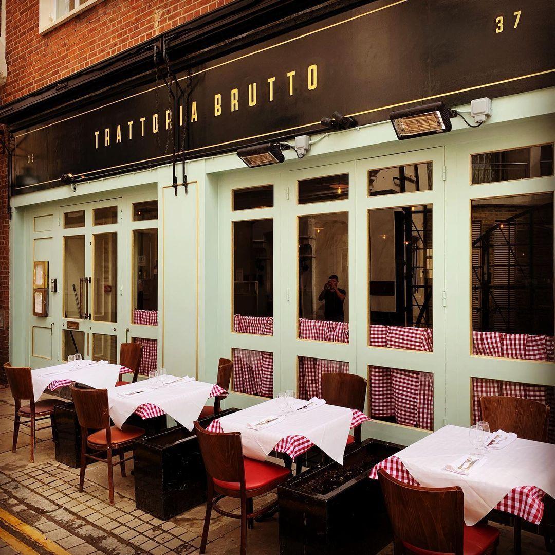 Street view of a quaint restaurant with a light green facade and a black sign reading Trattoria Brutto. Outdoor tables are set with red and white checkered tablecloths, covered with white sheets at at Brutto, Farringdon.