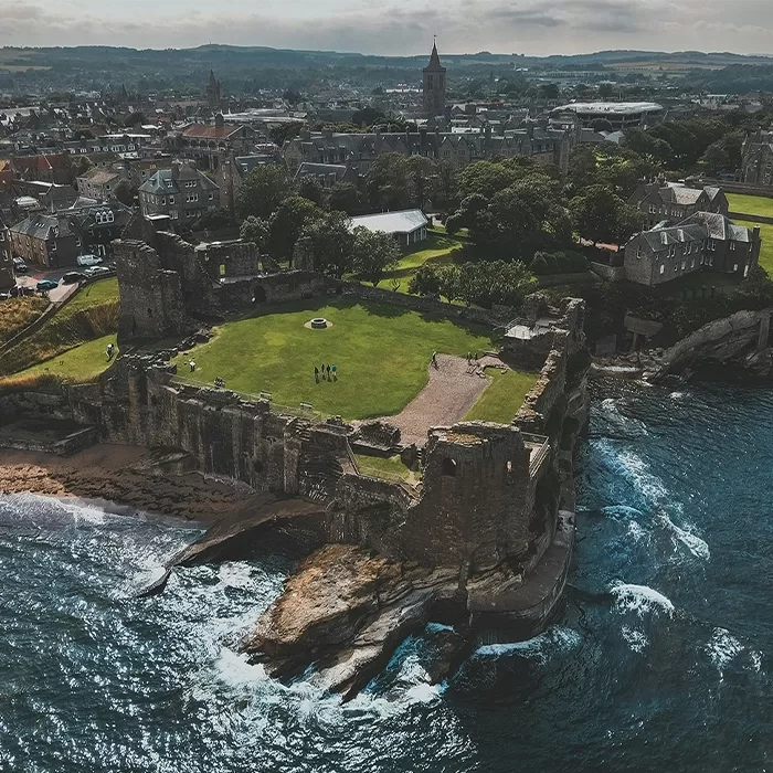 Aerial view of a historic stone castle ruin on a grassy cliffside overlooking the ocean, with a town and rolling hills visible in the background under a cloudy sky.