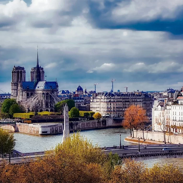 A scenic view of Paris with the Notre-Dame Cathedral on the left, the Seine River flowing through the city, and trees with autumn foliage under a partly cloudy sky.